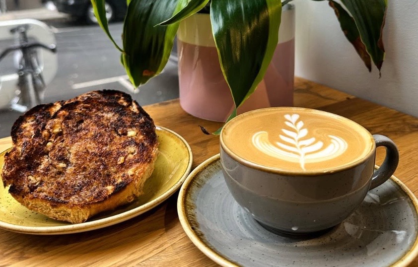 A latte with a fern pattern next to toasted bread on a wooden table